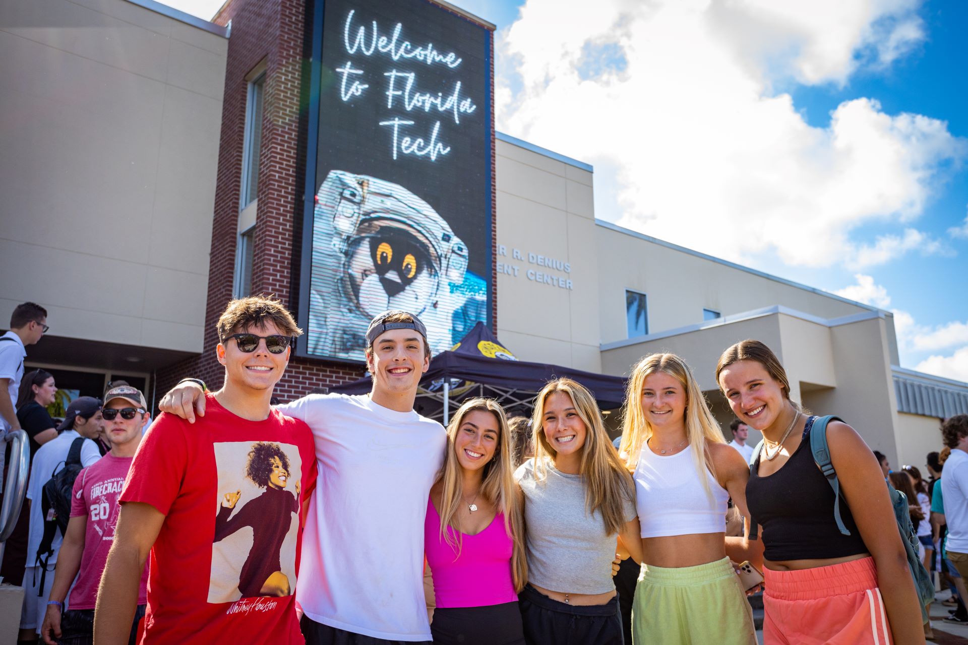 Students posing in front of the Panther Plaza at Florida Tech, with a welcome sign and the Clemente Center in the background, under a bright, clear sky